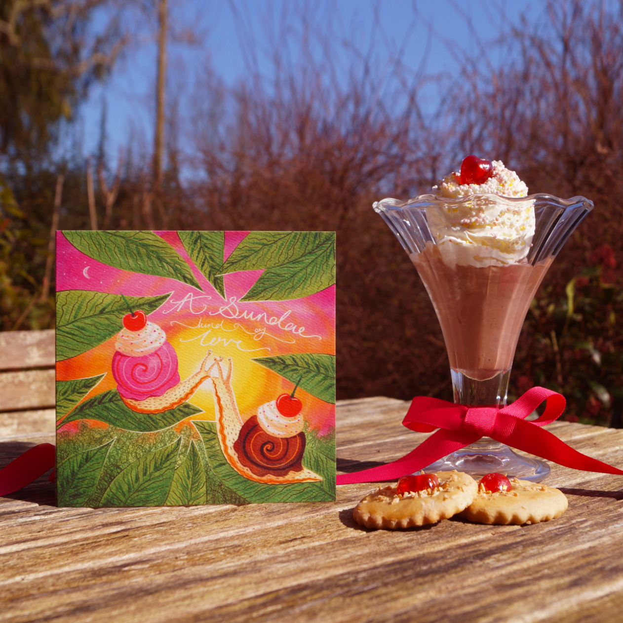 A Sundae Kind Of Love, Valentine's greetings card featuring a vibrant, romantic illustration of two snails with ice-cream and cherries on their shells, kissing in front of a sunset. Displayed standing next to an ice-cream sundae and biscuits on a wooden table outside. 14cm x 14cm, square in size and printed on premium quality 300gsm card stock. Designed by luxury gifts and stationery brand, Bella & The Moon