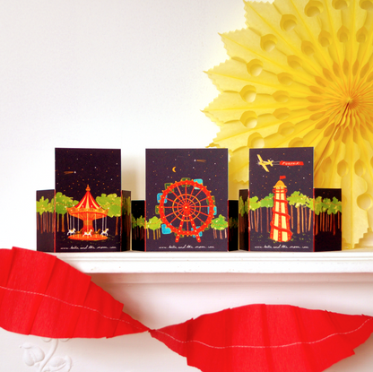 A decorative set of pop-up funfair cards with carnival-themed illustrations on a shelf with a yellow paper fan and a red garland. Kids crafts and gifts. Bella & The Moon gifts and stationery