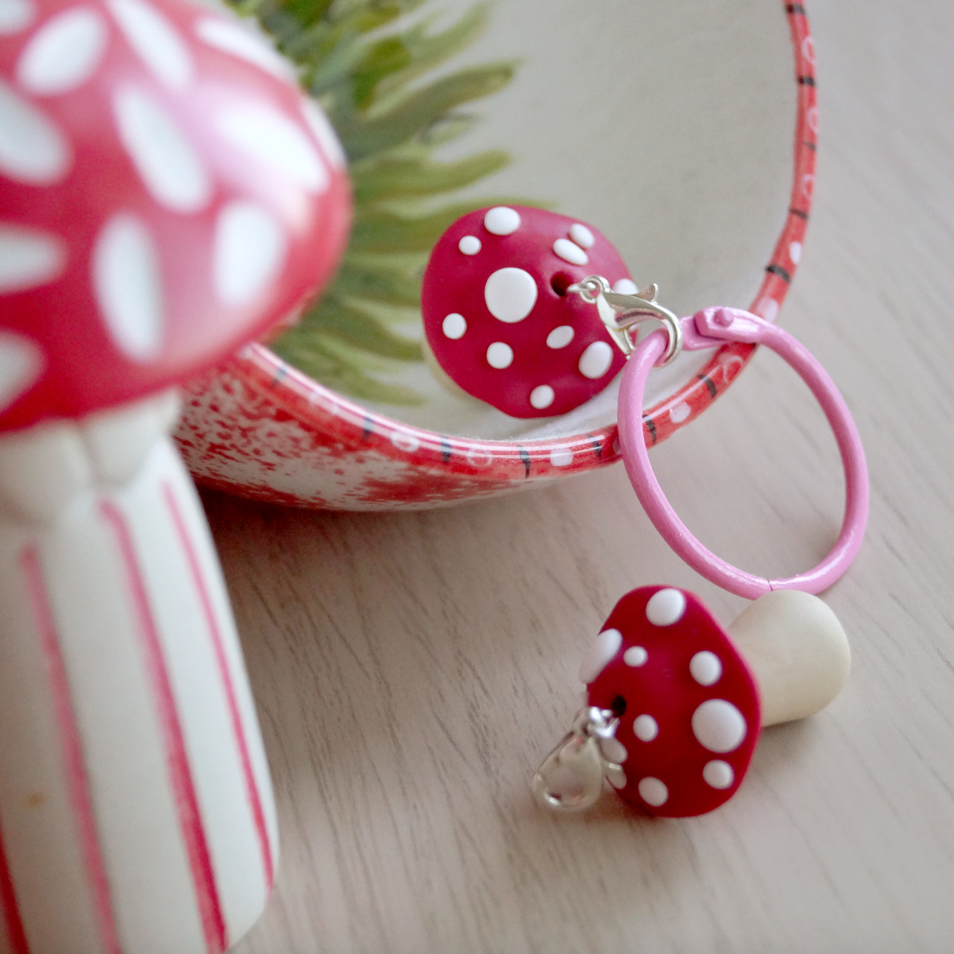A handmade polymer clay keyring in the shape of a red and white mushroom, displayed on a table with a decorative item in the background. Bella & The Moon gifts and stationery