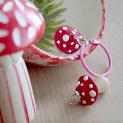 A handmade polymer clay keyring in the shape of a red and white mushroom, displayed on a table with a decorative item in the background. Bella & The Moon gifts and stationery