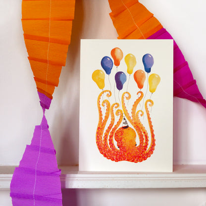Birthday or celebration greeting card featuring an orange octopus holding 8 colourful balloons. Displayed on a white shelf with orange and purple paper garlands. A5 in size and printed on 300gsm premium high quality card stock. Designed by luxury handmade gifts and stationery brand, Bella & The Moon.