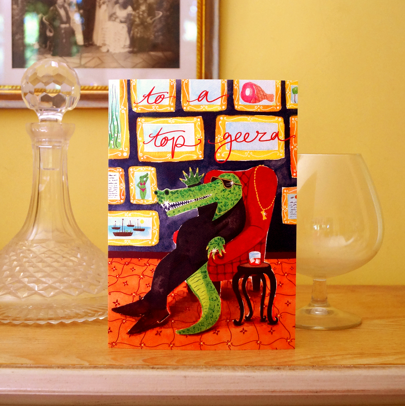 Greeting card with a crocodile lounging in a chair, surrounded by framed pictures, on a patterned background. The card stands on a cabinet with a brandy glass and wine carafe. A funny birthday, father's day or celebration card for men. A5 in size. Bella & The Moon gifts and stationery.