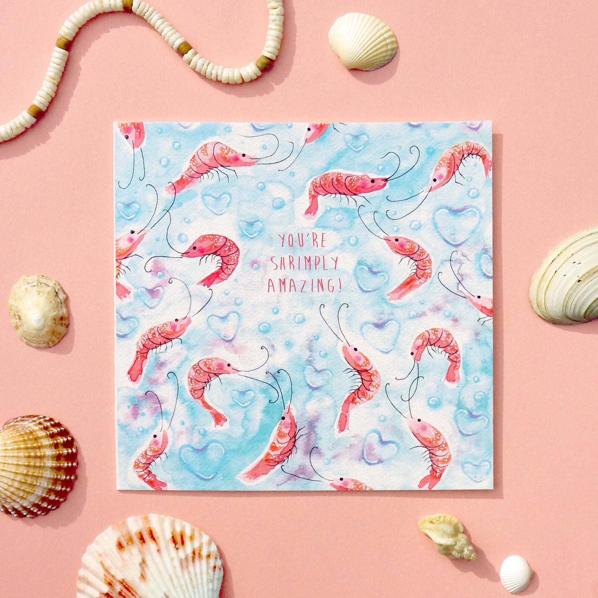 Love or valentine's day card with an underwater shrimp pattern and 'You're Simply Amazing' text on a light blue background with heart-shaped bubbles. Displayed on a pink table decorated in sea shells. 14cm x 14cm square in size and printed on premium quality 300gsm card stock. Designed by luxury handmade gifts and stationery brand Bella & The Moon