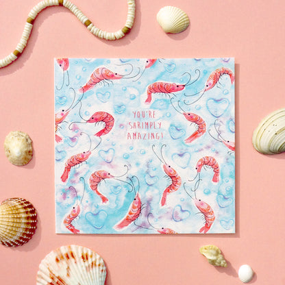 Love or valentine's day card with an underwater shrimp pattern and 'You're Simply Amazing' text on a light blue background with heart-shaped bubbles. Displayed on a pink table decorated in sea shells. 14cm x 14cm square in size and printed on premium quality 300gsm card stock. Designed by luxury handmade gifts and stationery brand Bella & The Moon