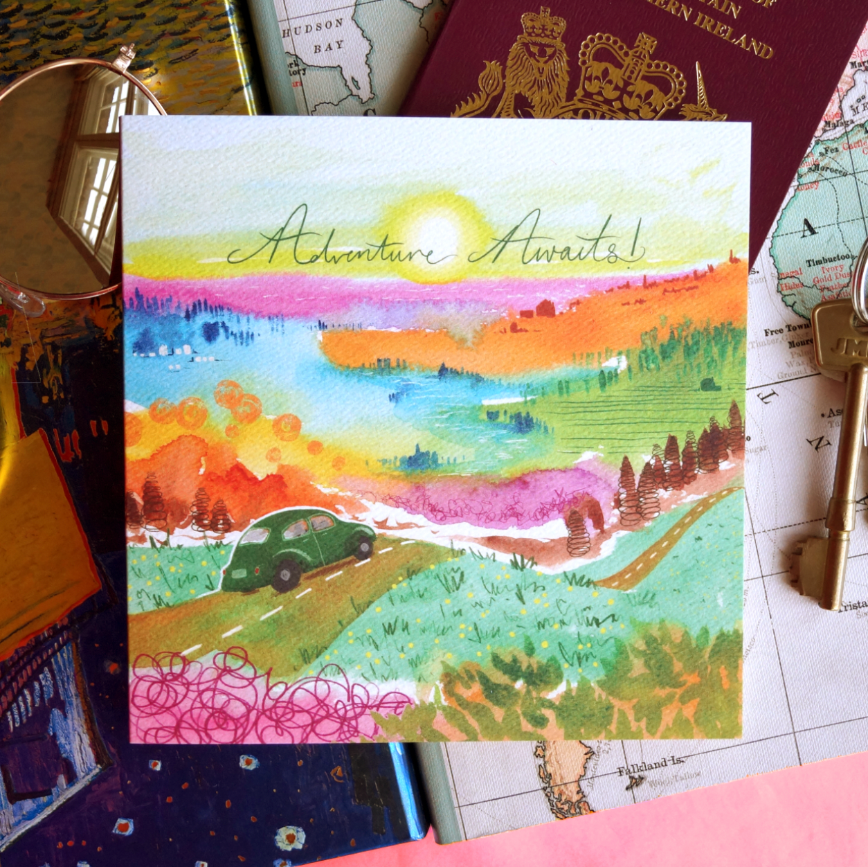 Adventure Awaits, traveller greetings card featuring a vibrant artistic landscape and a retro car. Displayed on a table with sunglasses, a passport, keys and a map. Perfect for travellers, backpackers, new drivers and exciting events. 14 x 14cm, square in size and printed on premium quality 300gsm card stock. Designed by luxury gifts and stationery brand, Bella & The Moon.