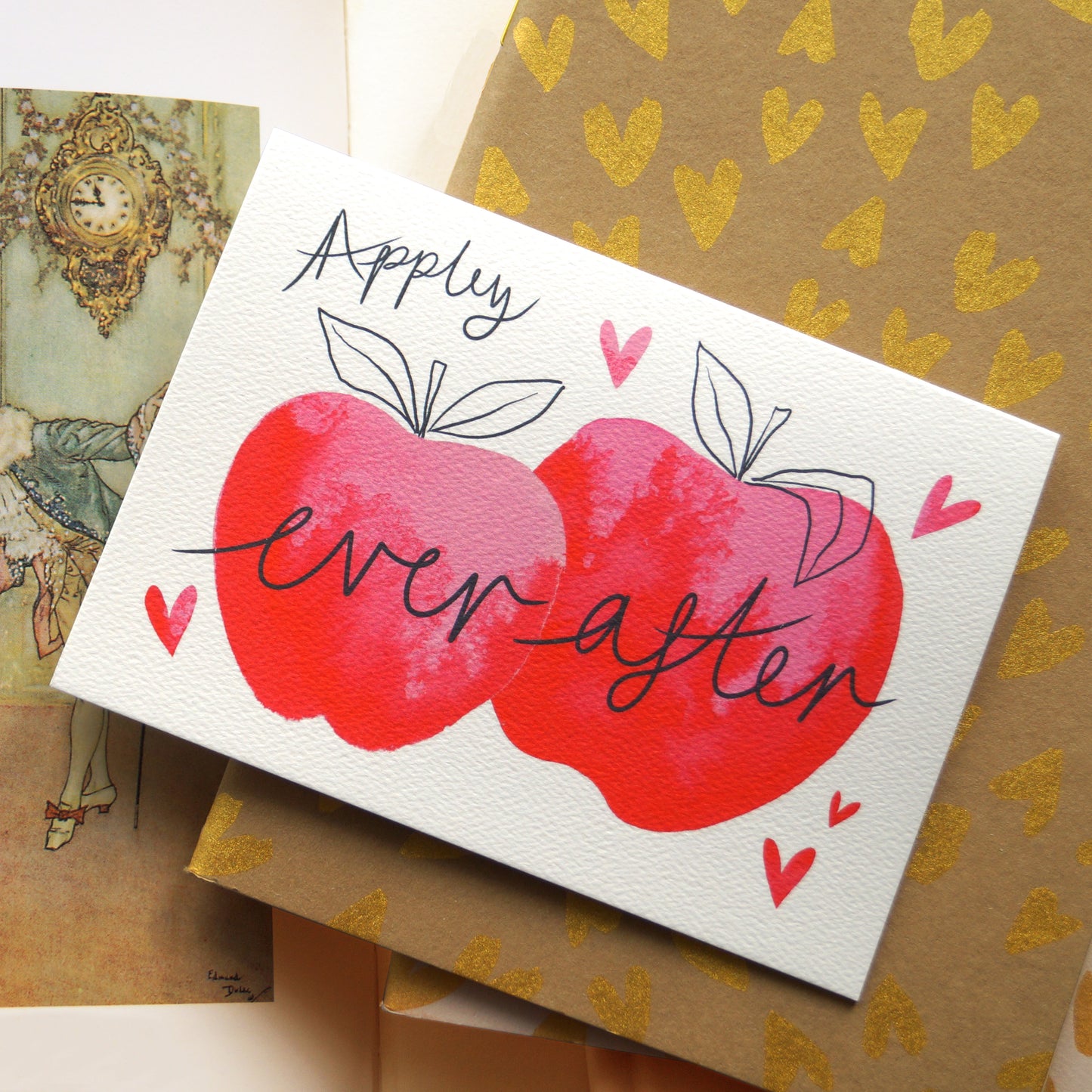 ‘Appley Ever After’ apple-themed greetings card featuring an apple related pun and two pink watercolour apples surrounded by love hearts. Displayed on top of gold embellished notebooks. Perfect for weddings, engagements, celebrating love, and Valentine's Day. A6 in size and printed on premium quality 300gsm card stock. Designed by luxury handmade gifts and stationery brand, Bella & The Moon.