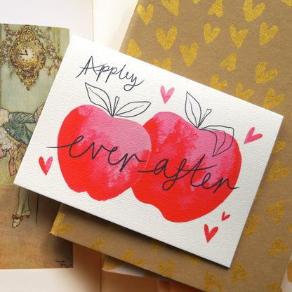 ‘Appley Ever After’ apple-themed greetings card featuring an apple related pun and two pink watercolour apples surrounded by love hearts. Displayed on top of gold embellished notebooks. Perfect for weddings, engagements, celebrating love, and Valentine's Day. A6 in size and printed on premium quality 300gsm card stock. Designed by luxury handmade gifts and stationery brand, Bella & The Moon.