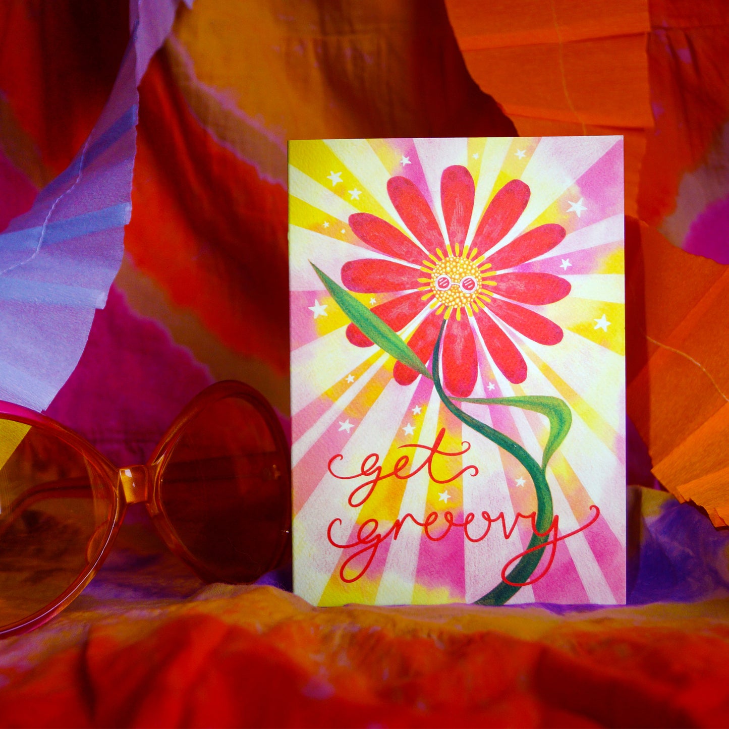 Get Groovy, retro, flower greetings card featuring a dancing flower with disco glasses and a hippie, tie dye, background. Displayed on orange tie-dye backdrop with big sunglasses and lilac paper garlands. Perfect for party animals, ravers, disco queens, hippies, celebrations, party invitations and birthdays. A6 in size and printed on premium quality 300gsm card stock. Designed by luxury gifts and stationery brand, Bella & The Moon.