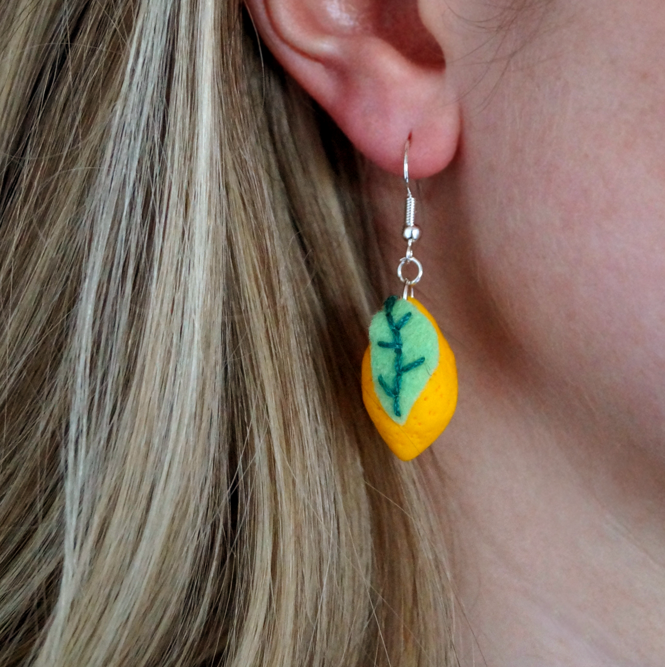 Handmade lemon-shaped earrings with green embroidered felt leaves, dangling on the earlobe of a model. Bella & The Moon gifts and stationery