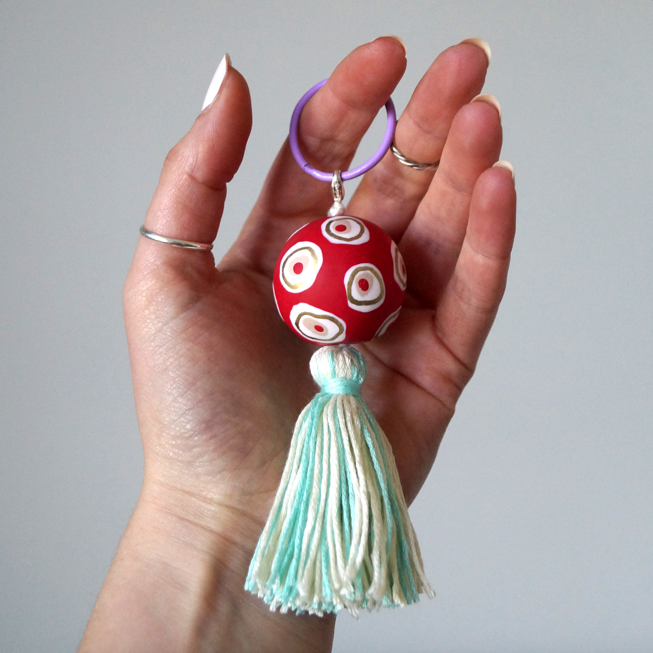 A hand modelling a handmade keyring featuring a red, gold and white handmade polymer bead and a handmade blue tassel. A keyring made to accessorise bags, keys and clothing. Bella & The Moon gifts and stationery