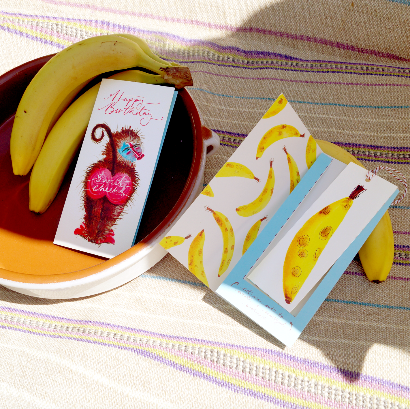 A funny baboon happy birthday greetings card with the words 'sweet cheeks'. It features a big-bottomed baboon on an extra special greeting card. The card includes a cut-out bookmark inside with a banana illustration. Displayed on a tablecloth with fresh bananas. DL in size. Bella & The Moon gifts and stationery 