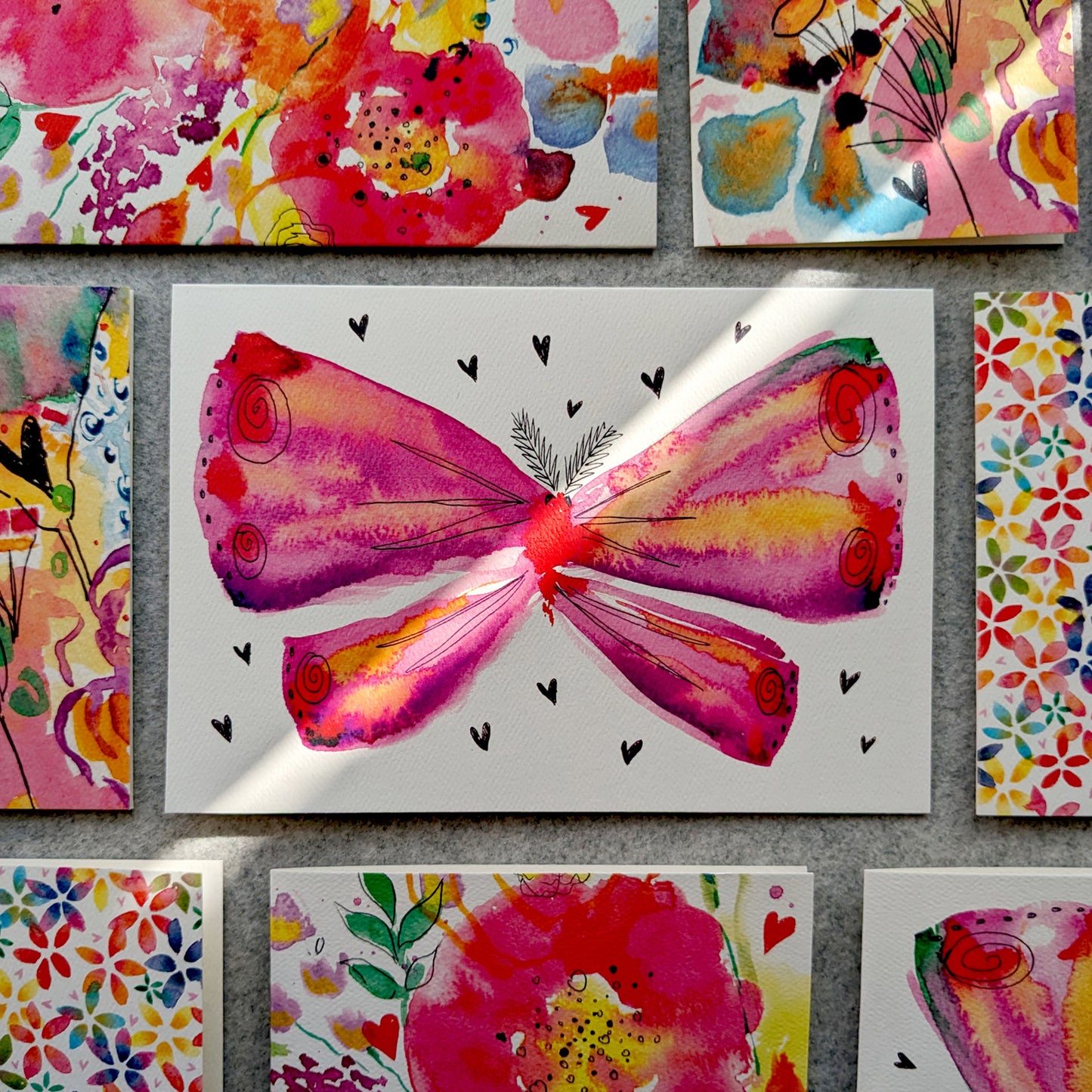 This Moth greetings card features a vibrant, pink, watercolour moth illustration surrounded by love hearts. Displayed on a grey felt table surrounded by other colourful cards. Perfect for christenings, baby showers, new mums, to say thank you, Mother's day, pink lovers, moth lovers or to celebrate love. A5 in size and printed on premium quality 300gsm card stock. Designed by luxury handmade gifts and stationery brand, Bella & The Moon.