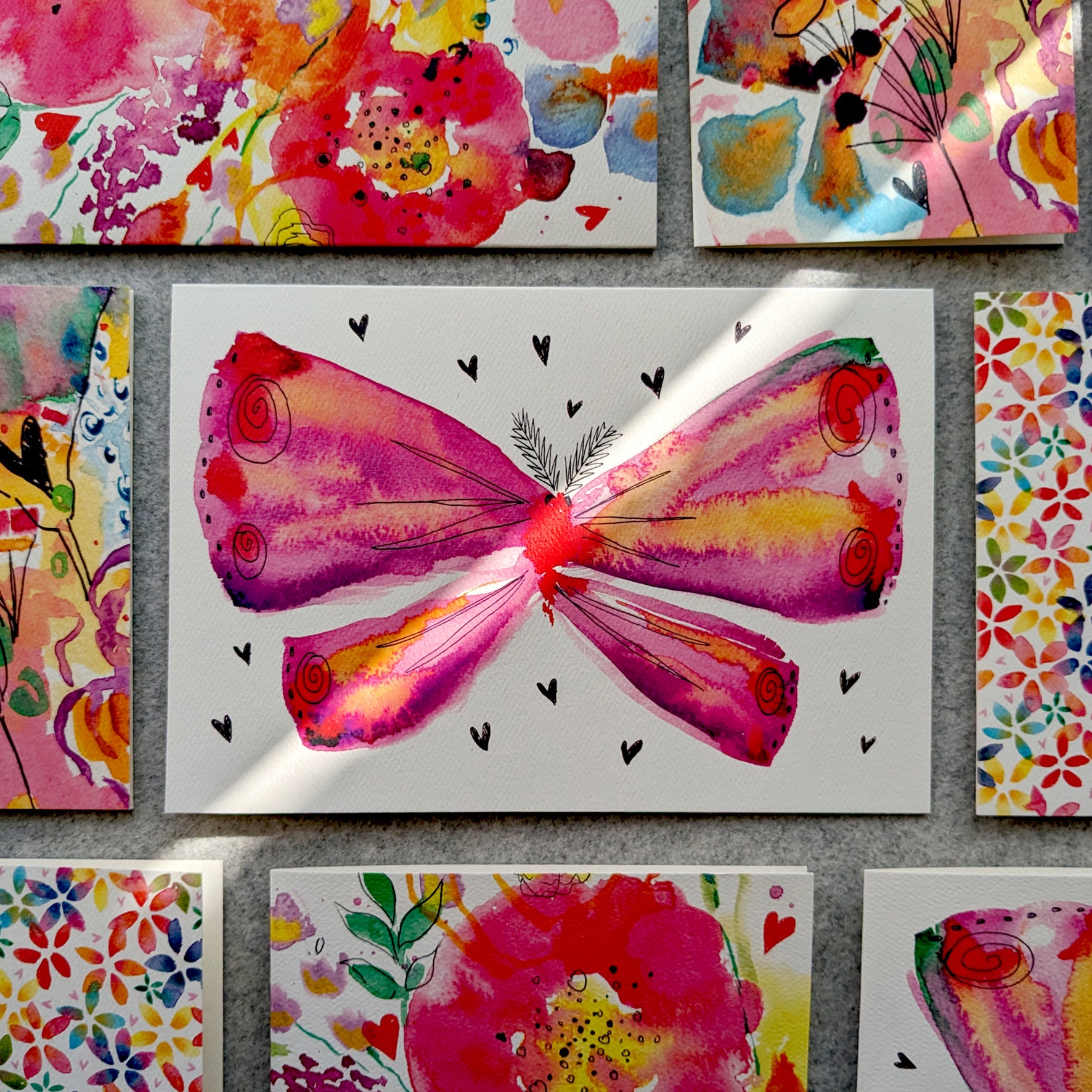 This Moth greetings card features a vibrant, pink, watercolour moth illustration surrounded by love hearts. Displayed on a grey felt table surrounded by other colourful cards. Perfect for christenings, baby showers, new mums, to say thank you, Mother's day, pink lovers, moth lovers or to celebrate love. A5 in size and printed on premium quality 300gsm card stock. Designed by luxury handmade gifts and stationery brand, Bella & The Moon.