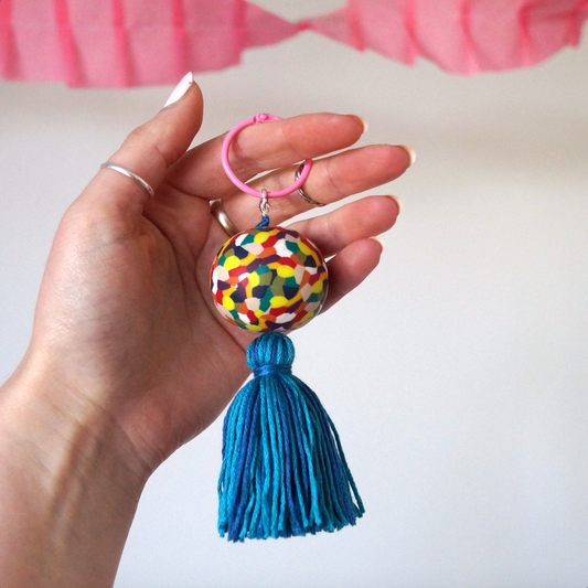 A hand modelling a handmade keyring featuring a colourful handmade polymer bead inspired by the children's elephant character 'Elmer'. It also features a handmade blue tassel. A keyring made to accessorise bags, keys and clothing. Bella & The Moon gifts and stationery