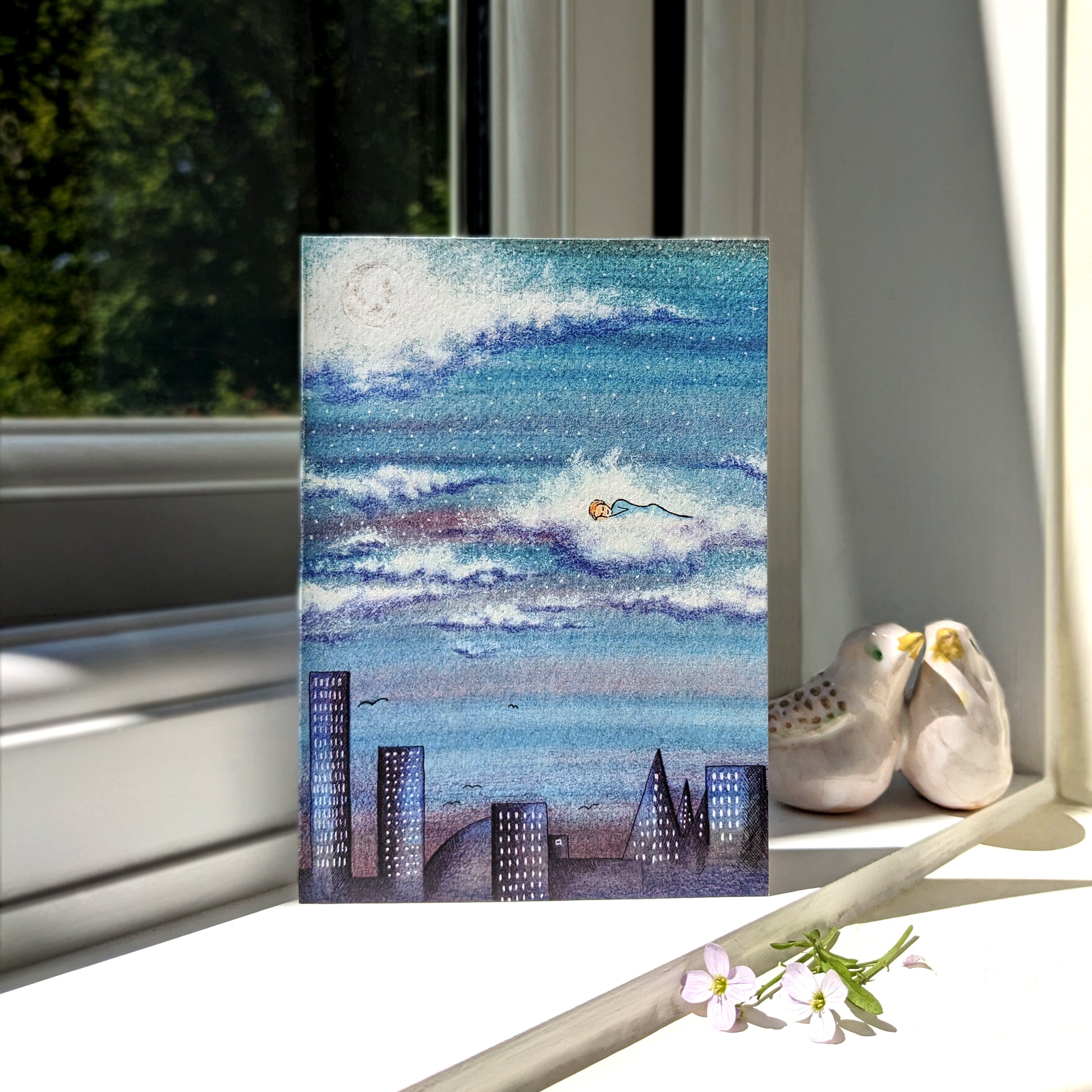 Sleeping in the Clouds greetings card featuring a person sleeping in a cloud above a city landscape, overlooked by the moon. Displayed on a sunny window cill with ceramic birds in the background. Perfect for birthdays, baby showers, or sending calming wishes. A6 in size and printed on premium quality 300gsm card stock. Designed by luxury handmade gifts and stationery brand, Bella & The Moon.