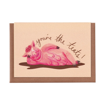 Funny greeting card featuring a pink pig wearing pearls and a nipple piercing, laying in a puddle of mud with text 'you're the teats!' on a pale pink background. A6 in size and printed on premium quality 300gsm card stock. Designed by luxury gifts and stationery brand, Bella & The Moon.